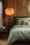 A Japandi-style bedroom with a low-profile dark bedframe, sage green linen bedding, neutral throw pillows, and a warm paper lantern floor lamp. A small side table holds ceramic vases and a floral arrangement, with wooden slat walls in the background.