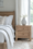 Minimalist bedroom with a wooden bed frame, crisp white bedding, a beige textured pillow, a wooden nightstand, and a ceramic vase with greenery near a bright window.