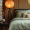 A Japandi-style bedroom with a low-profile dark bedframe, sage green linen bedding, neutral throw pillows, and a warm paper lantern floor lamp. A small side table holds ceramic vases and a floral arrangement, with wooden slat walls in the background.